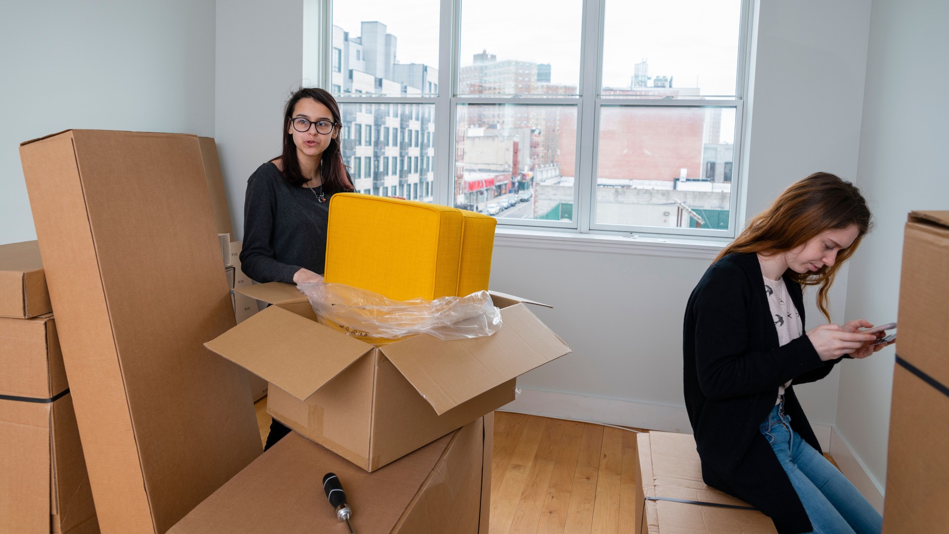 The family moving into the new apartment. The teenage girl unboxing and assembling furniture, when her older sister ordering pizza with her smartphone.